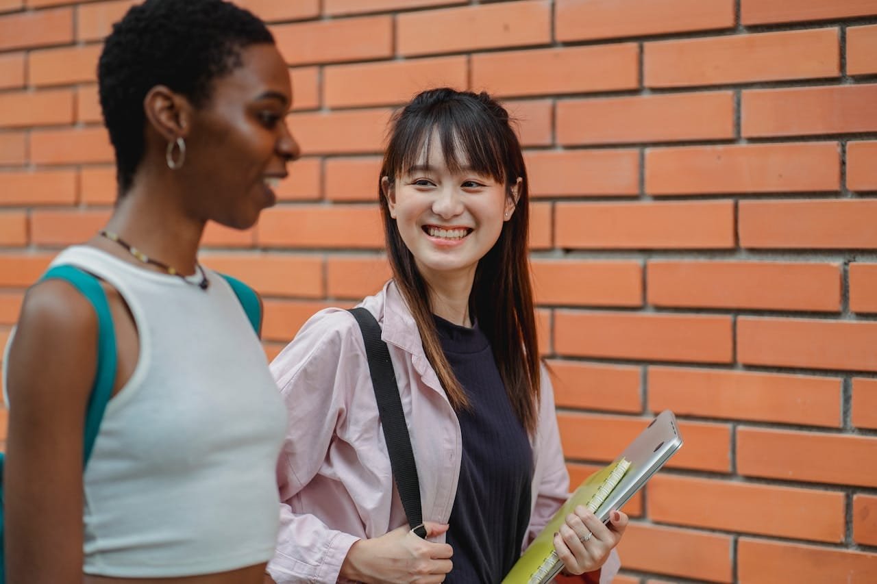 Two diverse female students conversing cheerfully outdoors on campus.