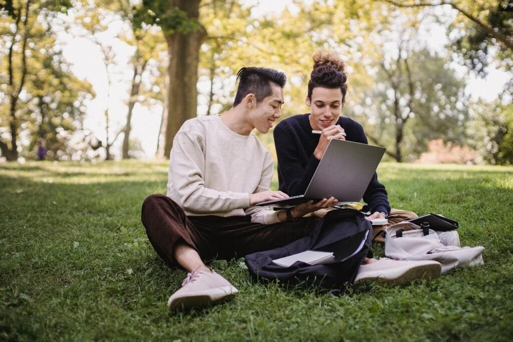 Two young men studying on a laptop while sitting on grass in a park.