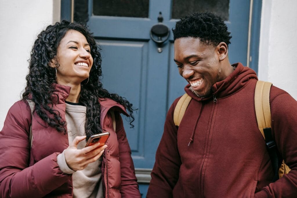 Two young adults smile and chat in front of a blue door, enjoying a light moment outdoors.